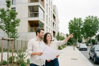 Couple d'acheteurs devant un immeuble neuf à Bagneux, tenant des plans d'architecte et souriant